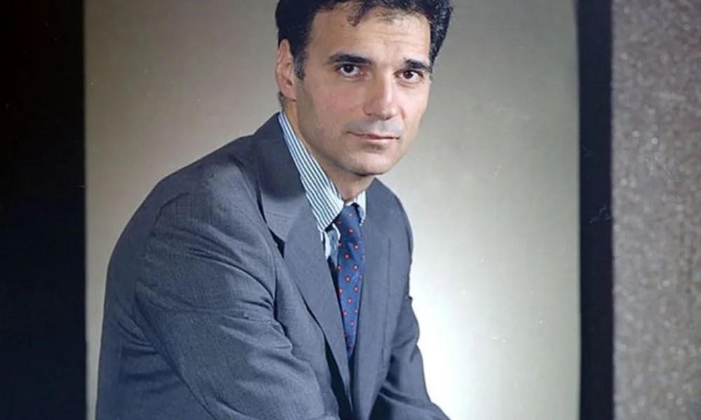 A man in a suit and tie sits against a neutral background