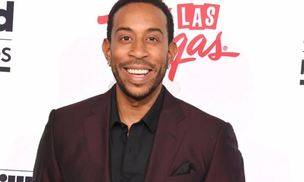 Smiling man in a dark suit stands against a backdrop-luda net worth