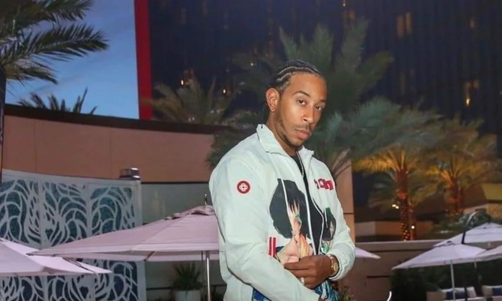 Man in a colorful jacket with braided hair stands confidently outdoors at dusk-luda net worth