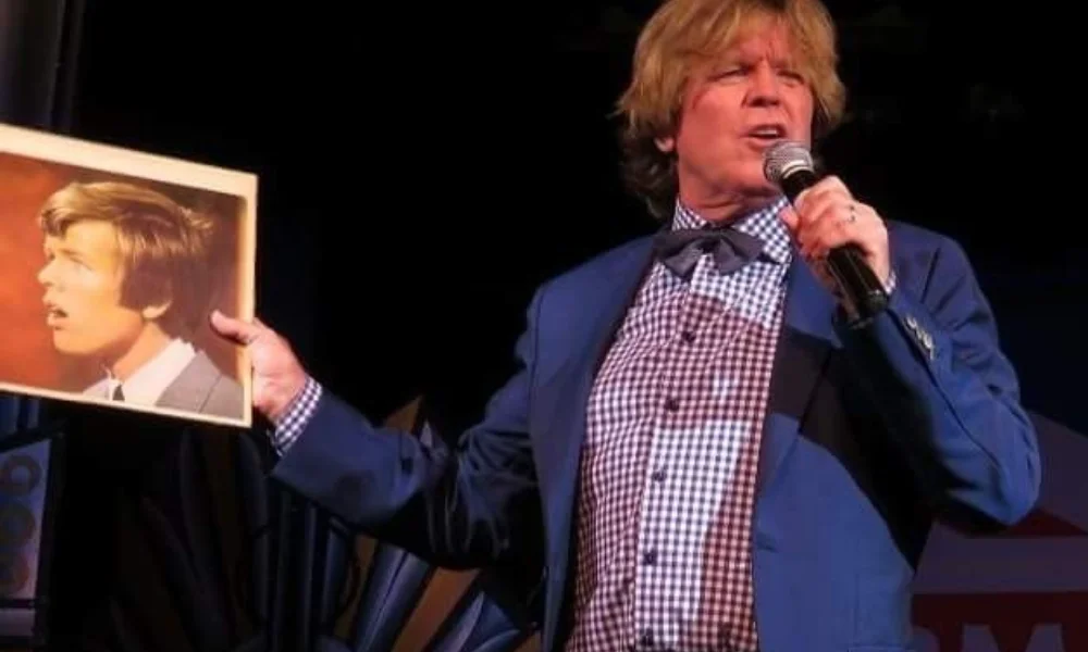 A man in a blue suit and checkered shirt speaks into a microphone